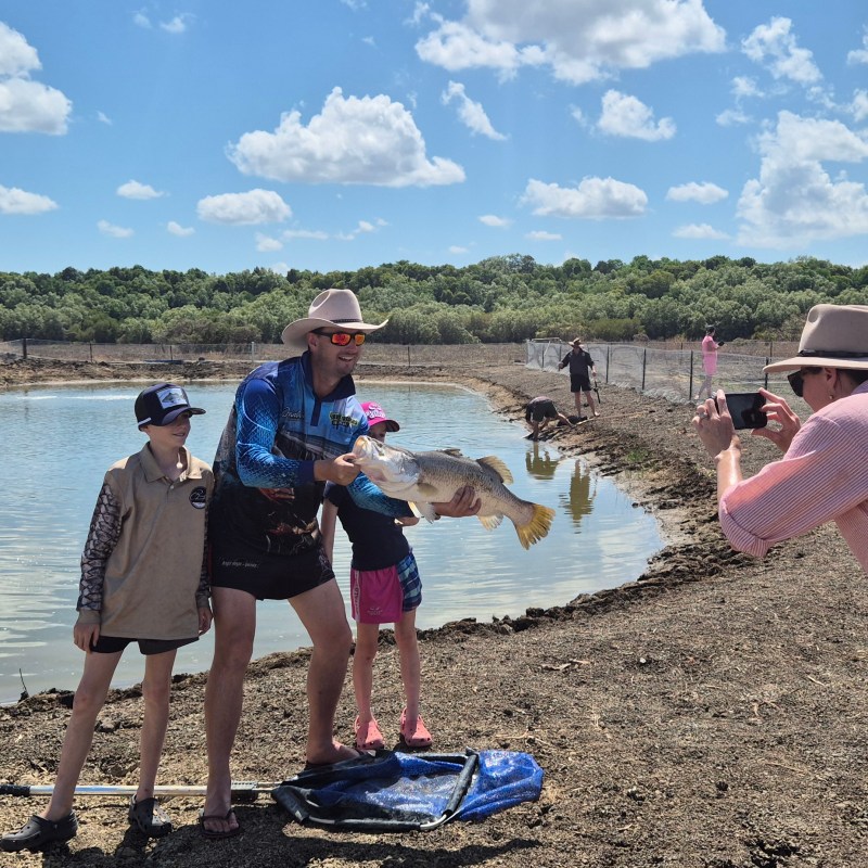 Family by a pond, man holding large fish, photo being taken, sunny day.