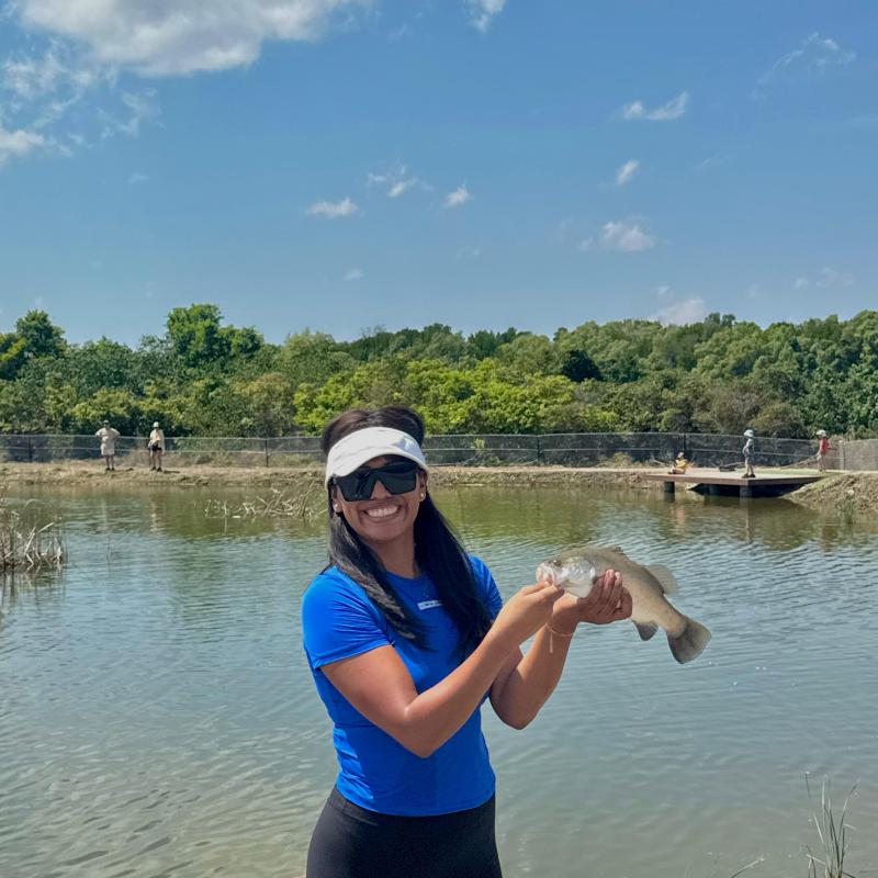 Woman in a blue shirt holding a fish on a dock by a pond, with trees and a blue sky in the background.
