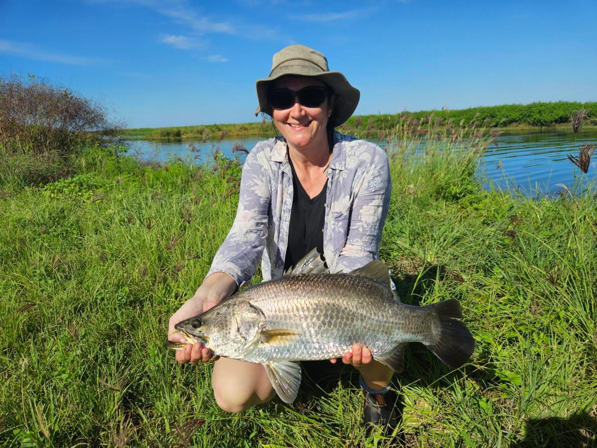 Person with hat and sunglasses holding a large fish by a grassy riverbank.