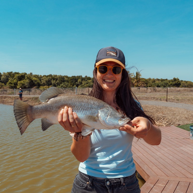 Person holding a fish on a dock by a body of water under a clear blue sky.