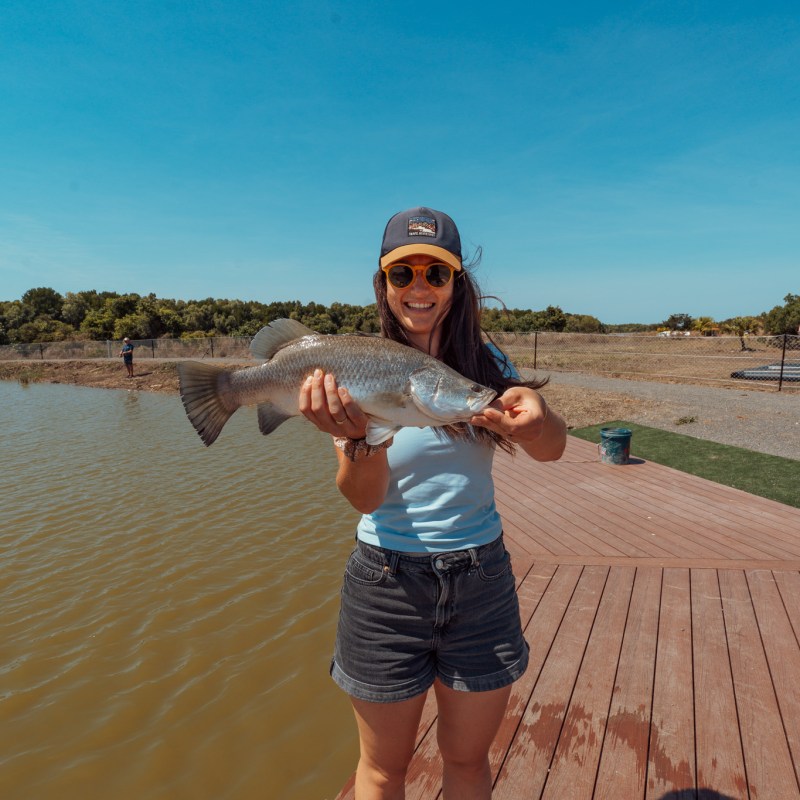 Woman on dock holding a fish, wearing a cap and sunglasses, with water and trees in the background.